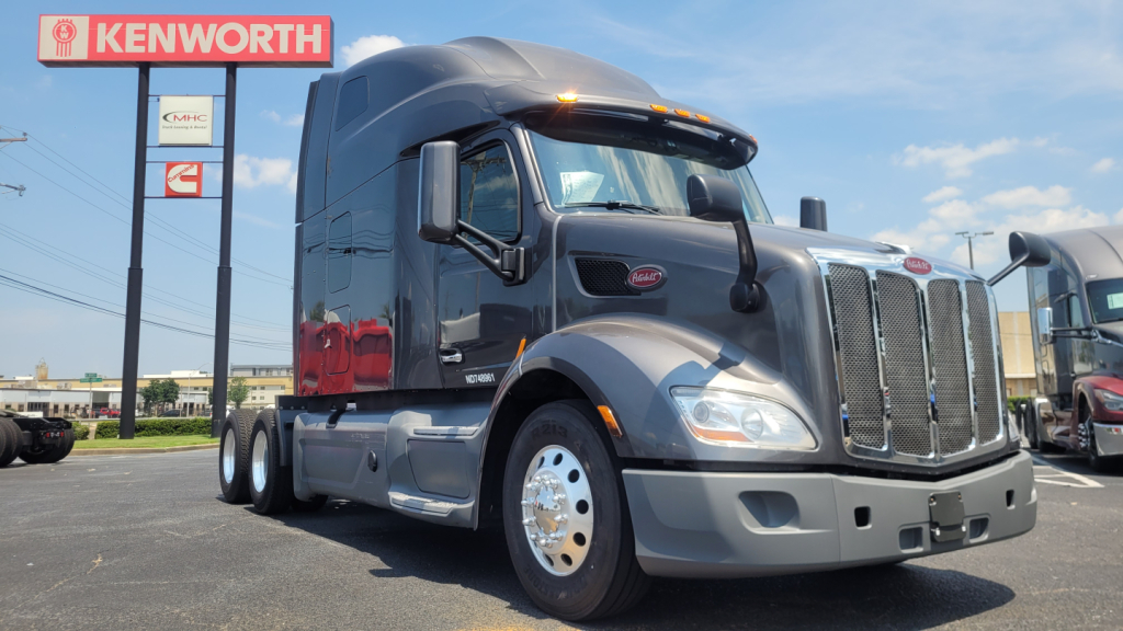 Peterbilt 579 conventional sleeper tractor in a used truck lot with Kenworth sign in background. Visible text on truck includes Peterbilt logo on grille and chrome bumper; no detailed specs readable.