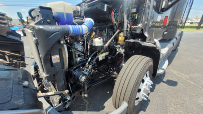 Peterbilt 579 engine bay view with exposed components and front wheel, showing hoses, wiring, and radiator area.