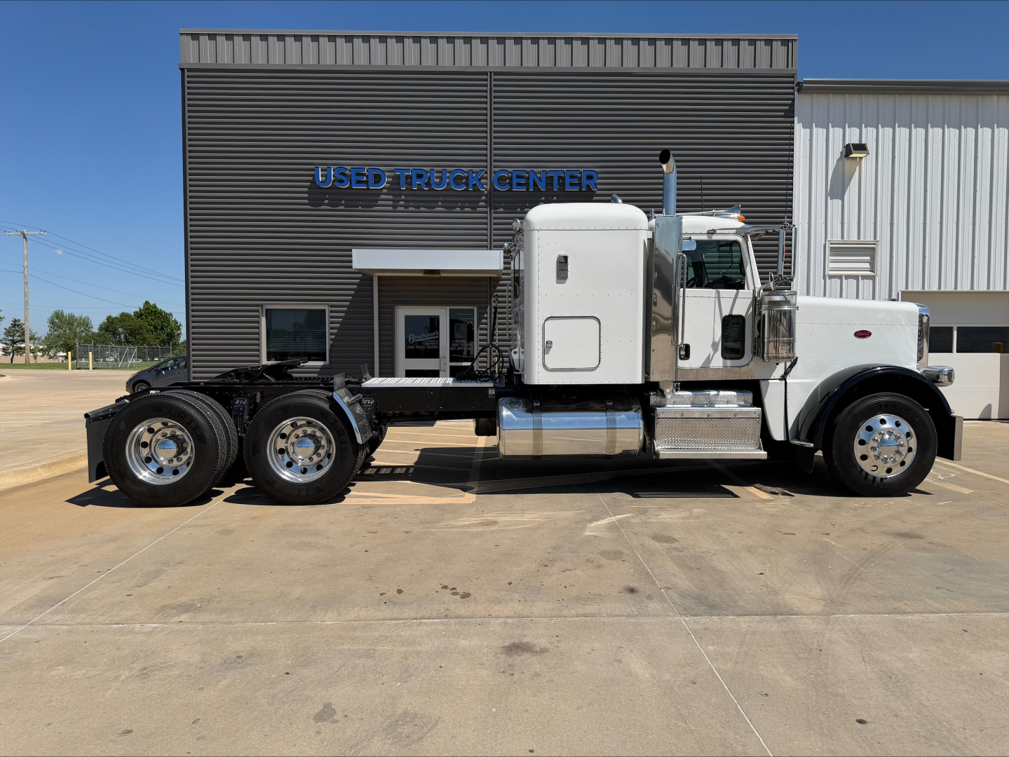 Peterbilt 389 conventional sleeper in white with chrome exhaust and dual rear axles, side view parked in front of building labeled Used Truck Center.