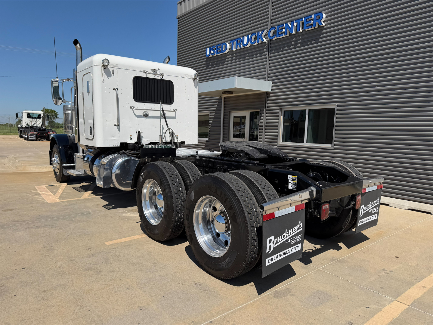 2015 Peterbilt 389 tractor with white cab, black chassis, dual rear axles, and rear mud flaps. Building with USED TRUCK CENTER sign in background. Visible text on rear mud flaps and license plate area.