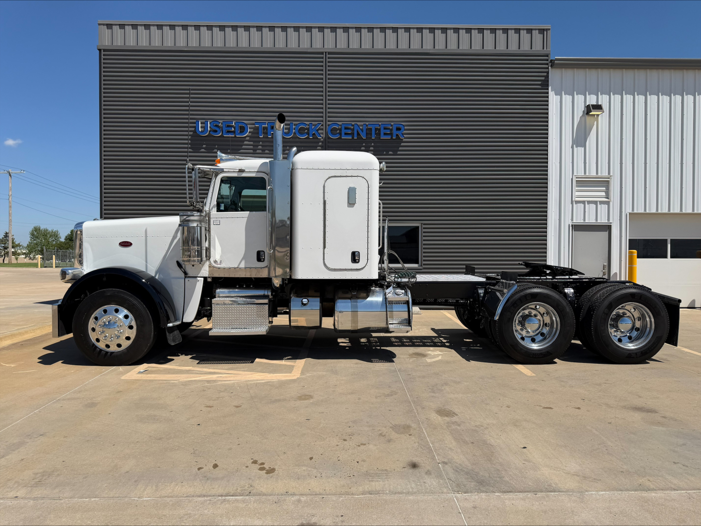 Peterbilt 389 conventional sleeper truck parked in lot with chrome exhaust stacks and aerodynamic cab; side view shows dual rear axles and gasoline/diesel tanks.