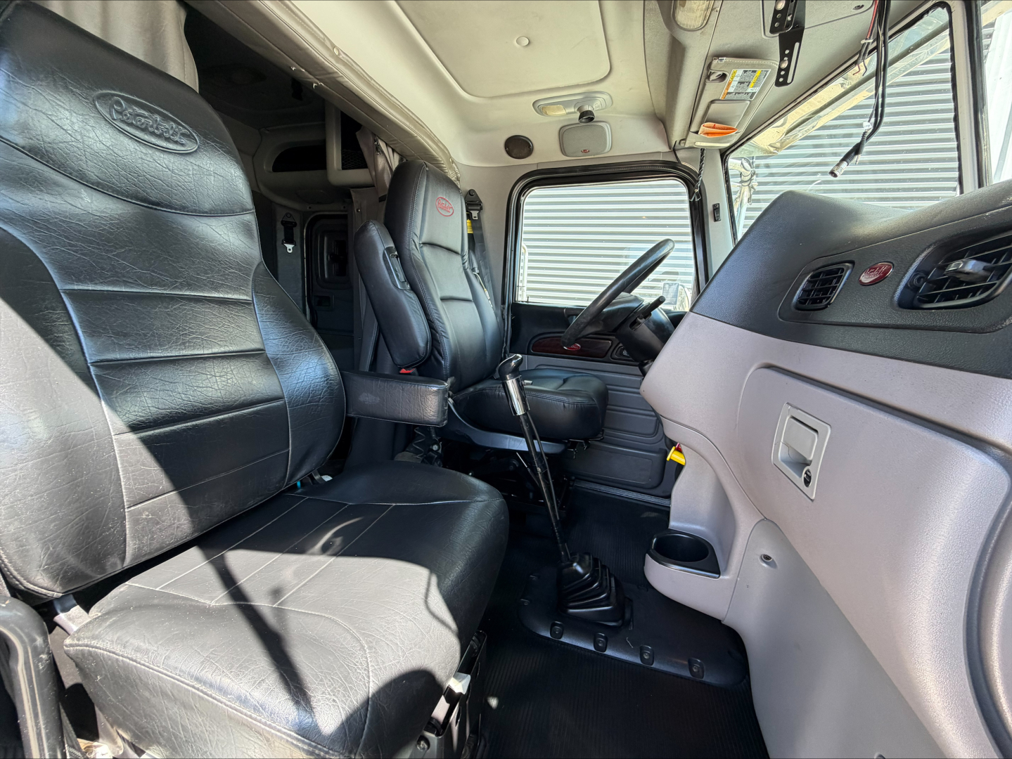 Peterbilt 389 interior cab view with two black leather seats, manual gear shifter, steering wheel, and dashboard; visible Peterbilt logo on seat; light gray interior panels.