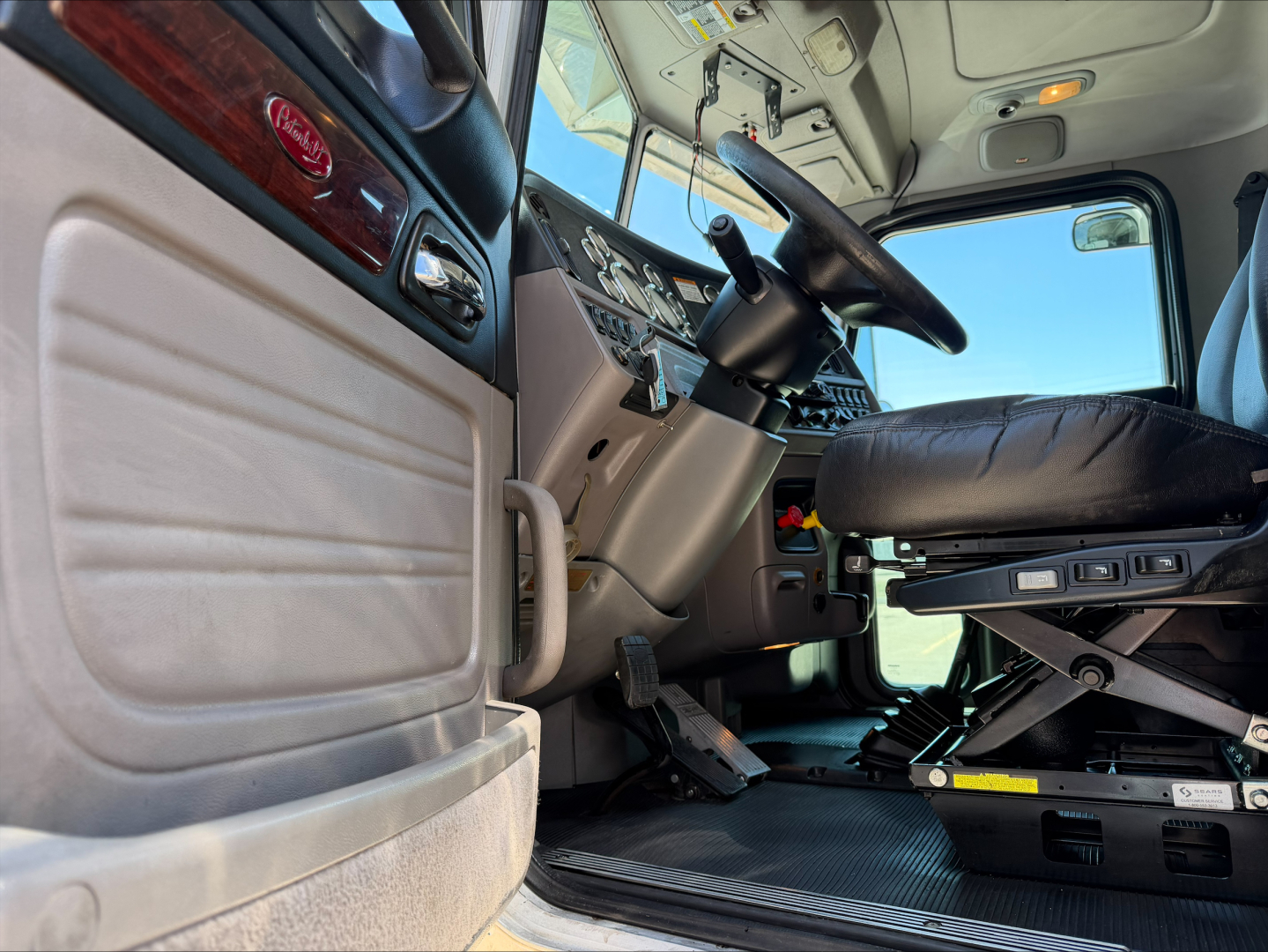 Interior of a Peterbilt 389 cab showing steering wheel, driver seat, pedals, and dashboard from a low angle.