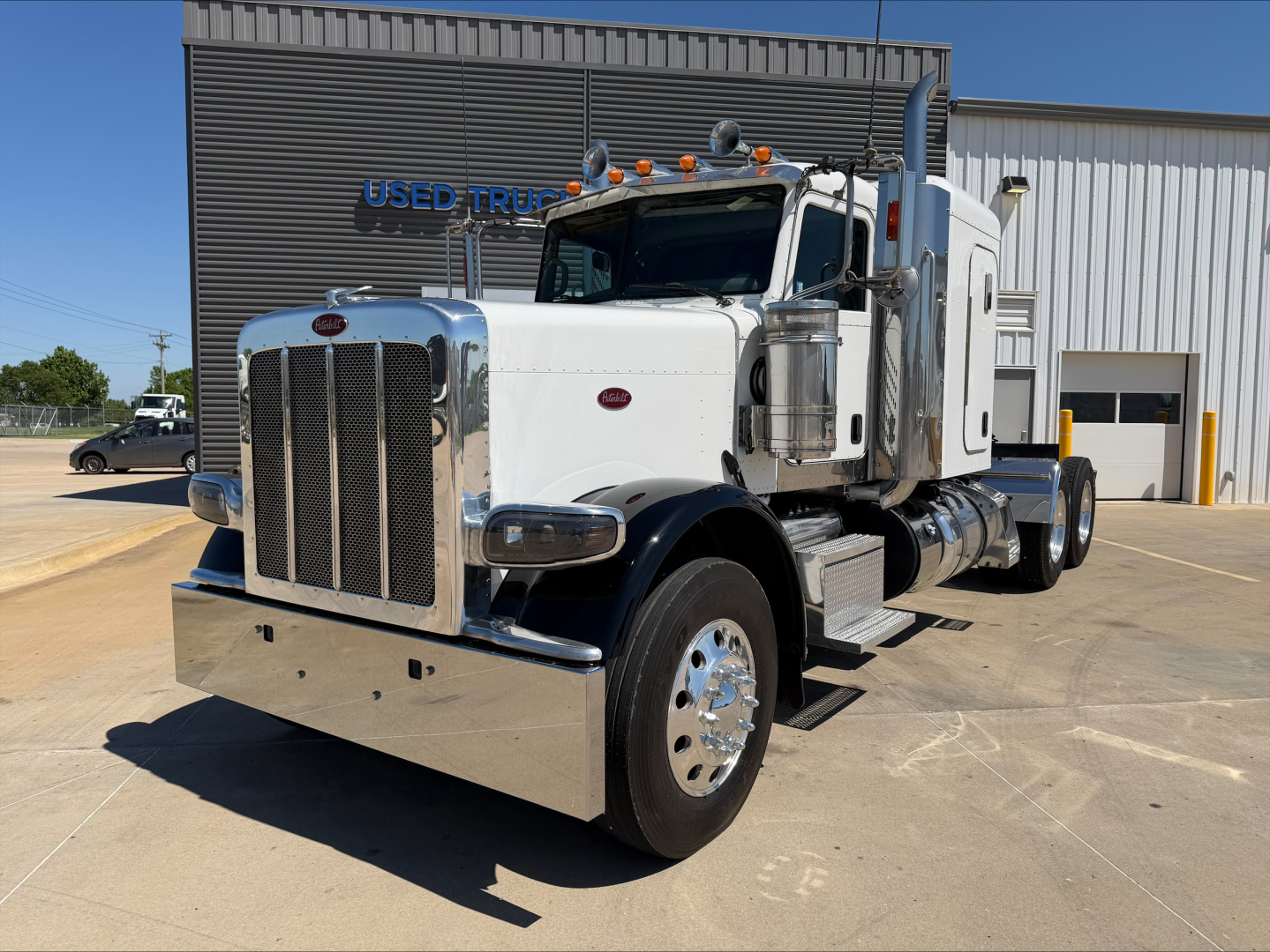 Peterbilt 389 heavy duty truck in front of dealership, chrome grille, white cab, large chrome bumper and exhaust stacks visible.