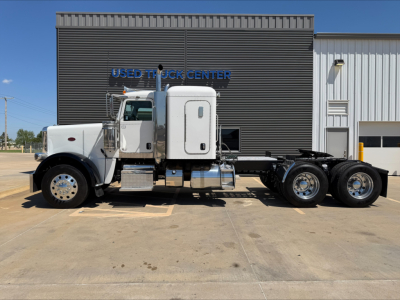 Peterbilt 389 conventional sleeper truck parked in lot with chrome exhaust stacks and aerodynamic cab; side view shows dual rear axles and gasoline/diesel tanks.