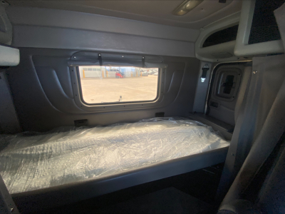 Truck cab sleeper area of a 2015 Peterbilt 389 showing a single bed with plastic cover, side window, overhead storage, and cabinet door.