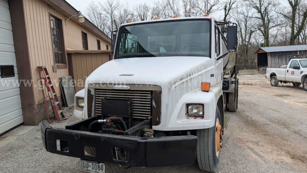 Truck front view of a white 1999 Freightliner FL70 with a black front bumper, grille, and damaged area; orange reflectors on the fenders; stock yard in background.