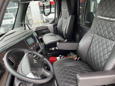 Interior of a Western Star truck cab with diamond-stitched seats, steering wheel, and dashboard; visible are control panel, central touchscreen, and gear selector. Passenger seat shows Western Star logo on headrest.
