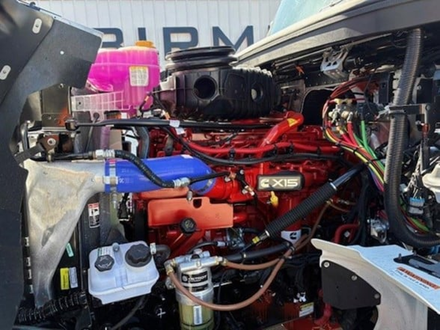Truck engine bay with red CX15 engine and various hoses and wiring visible; blue coolant hose, white reservoir, and metal components in foreground.