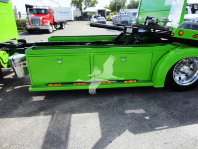 Green Peterbilt 337 rollback truck side panel with storage boxes and orange marker lights, part of a recreational equipment in a dealership lot.
