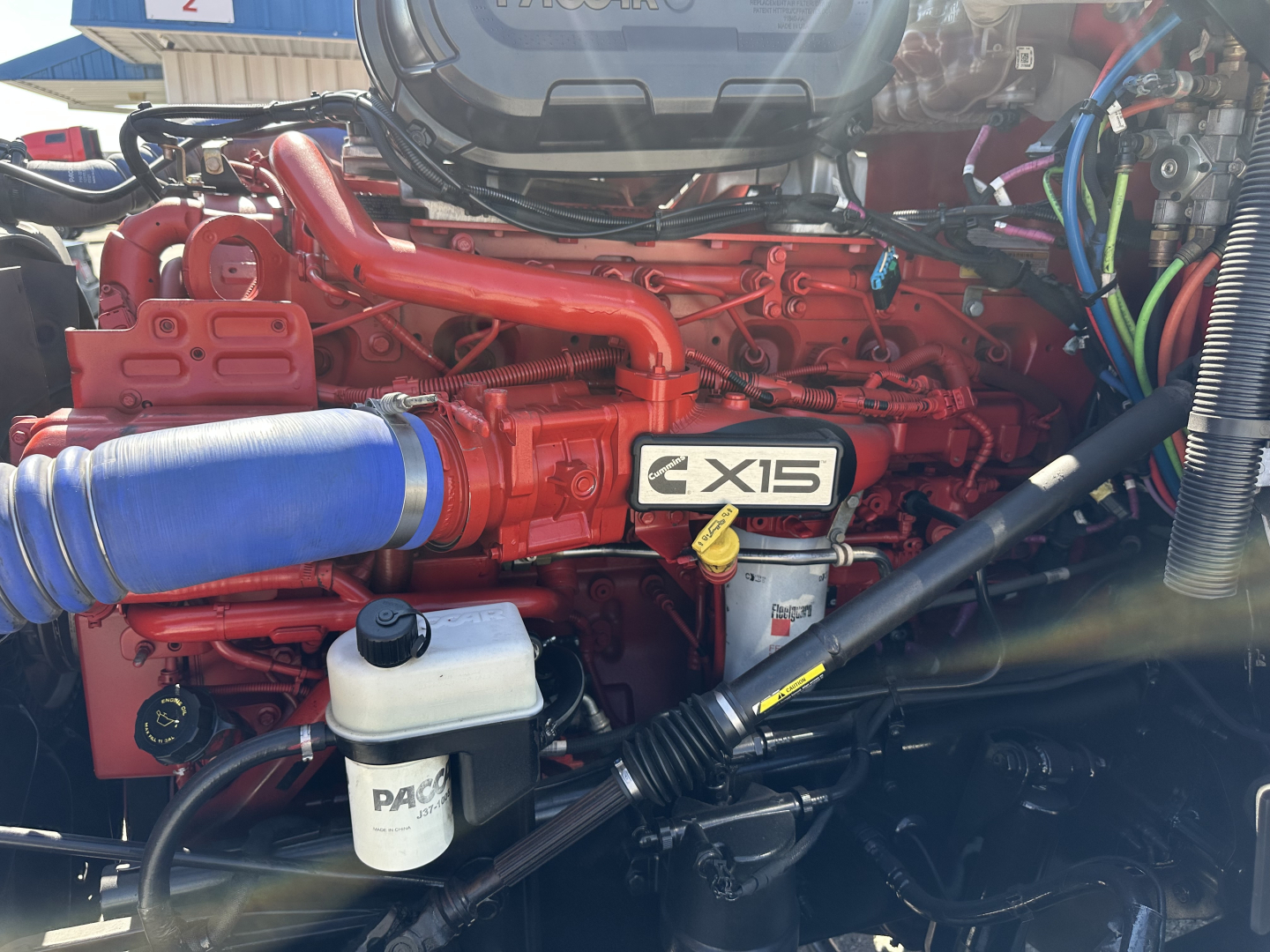 Peterbilt 579 engine bay showing CX15 diesel engine with red intake manifold, blue hose, white PAG filter canister, and surrounding hoses and wiring.