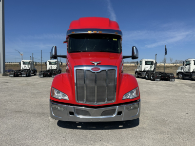 Peterbilt 579 front view showing grille, headlights, and chrome bumper; red cab with black windshield, tag lines, and side mirrors; truck lot in background.