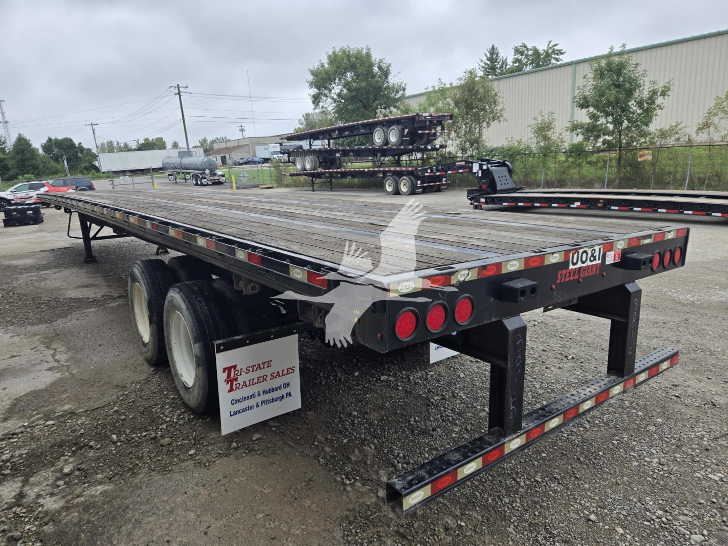 trailer flatbed showing steel deck and dual rear ramps with taillights; visible signage reads 0081 STEEL TRAILERS; yellow caution tape on edge; gravel lot background