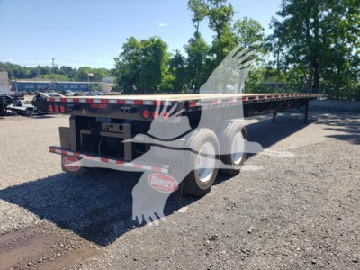 Flatbed trailer showing dual rear axles and long deck with reflective tape along edges.