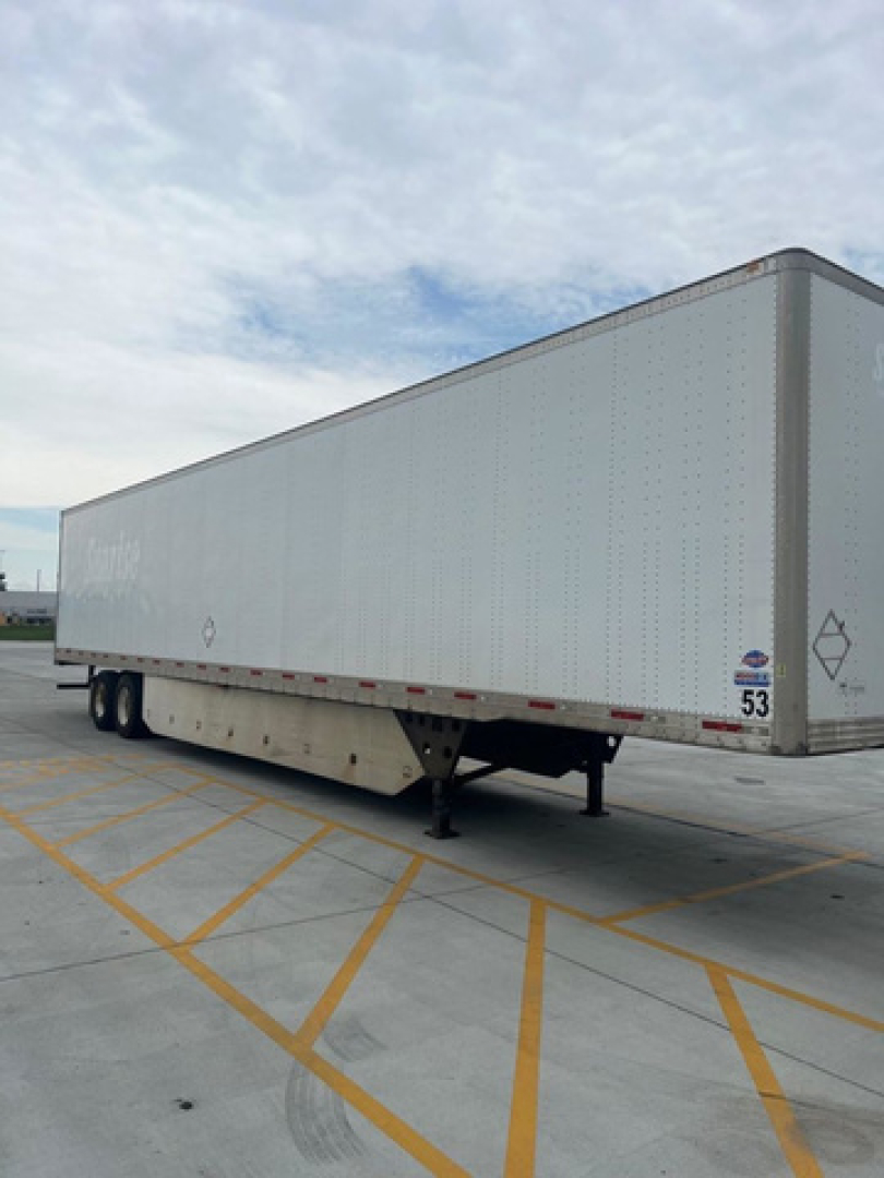 Trailer van side view showing utility box truck trailer with raised landing gear and tandem axles, white exterior with small diamond logo near front; parking lot lines visible.