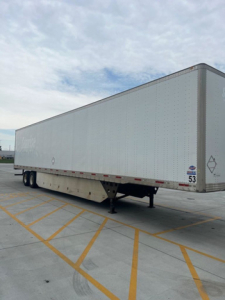 Trailer van side view showing utility box truck trailer with raised landing gear and tandem axles, white exterior with small diamond logo near front; parking lot lines visible.