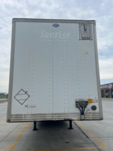 Rear of a white utility van trailer with a small logo near top, a black chute, and access doors; cargo area showing riveted panels and stabilizing legs under trailer