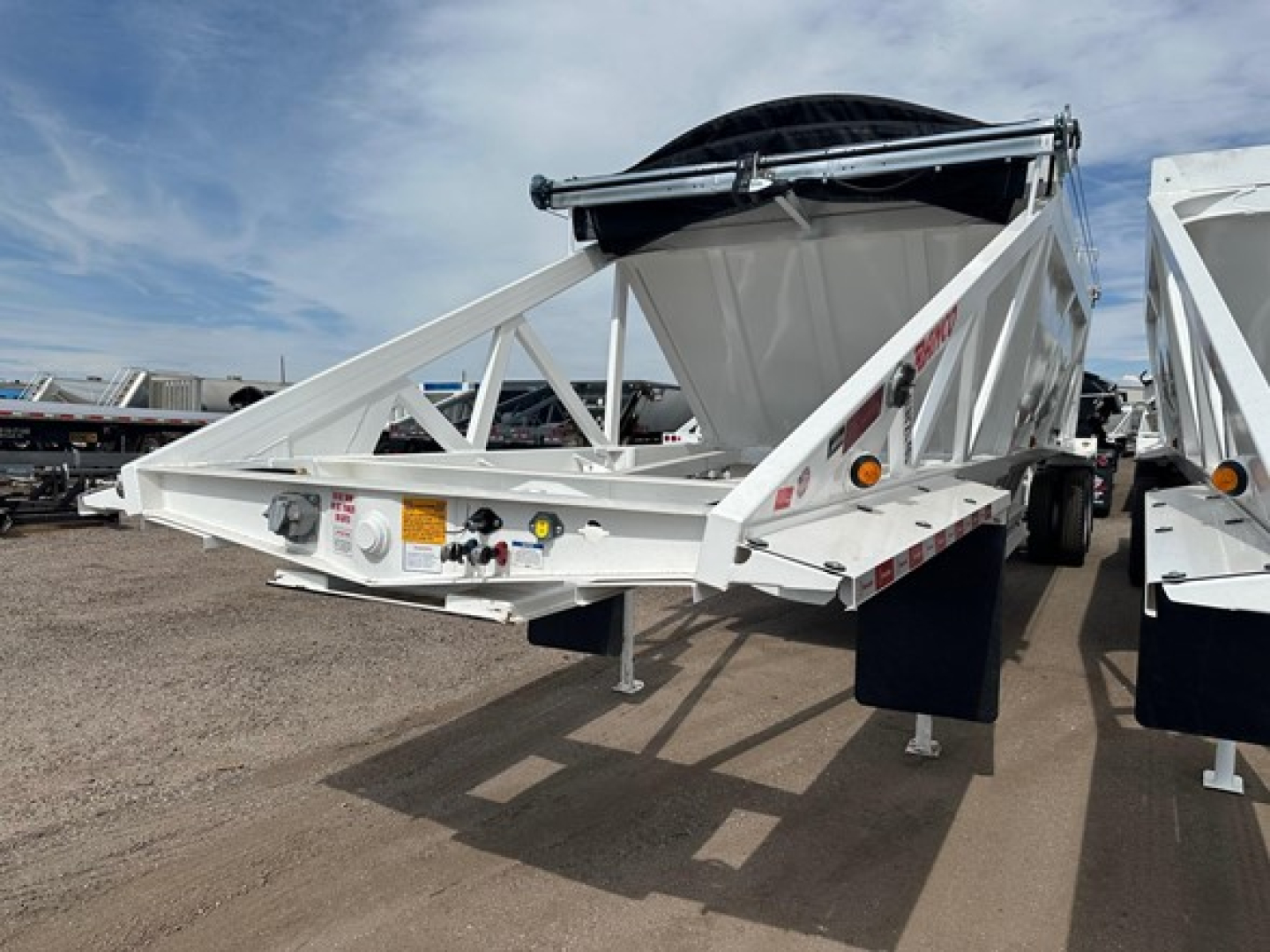 Trailer front section showing Ranco air ride bottom dump trailer with triangular frame, white paint, spare tire mounted on top, safety chains, labels and hydraulic components visible on the front rail.