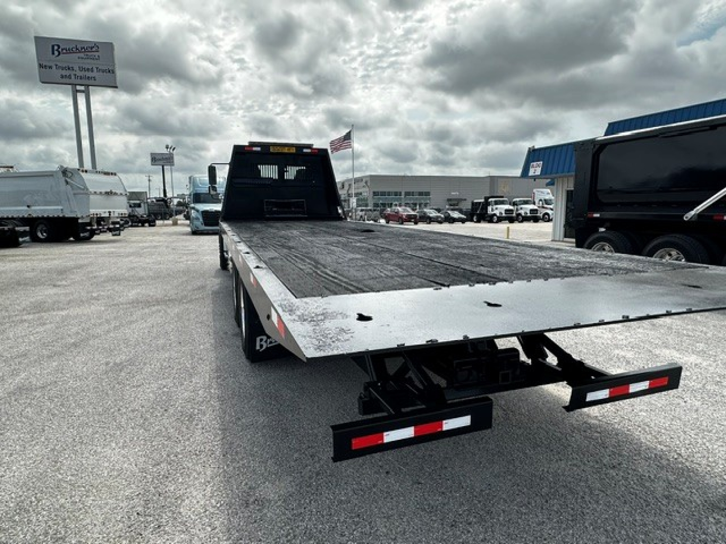 Flatbed rollback truck bed with black deck and rear taillights visible, Freightliner M2106-style equipment in a dealership lot.