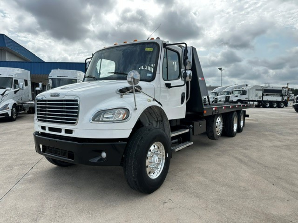 Freightliner M2106 rollback tow truck in white cab with black dump bed, front grille, round side mirror, and dual rear axles parked in lot