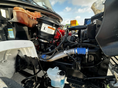 Freightliner M2106 engine bay with visible components including coolant reservoir, hoses, and engine block. Includes a label on a circular cap and blue hose clamps.