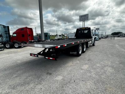 Flatbed tow truck with triple rear axles and a black ramp bed; rear bumper lights; small sign on pole in distance; overcast sky.