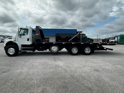 Freightliner M2106 rollback tow truck side view showing long flatbed with black bed and white cab, dual rear axles, and chrome wheels.
