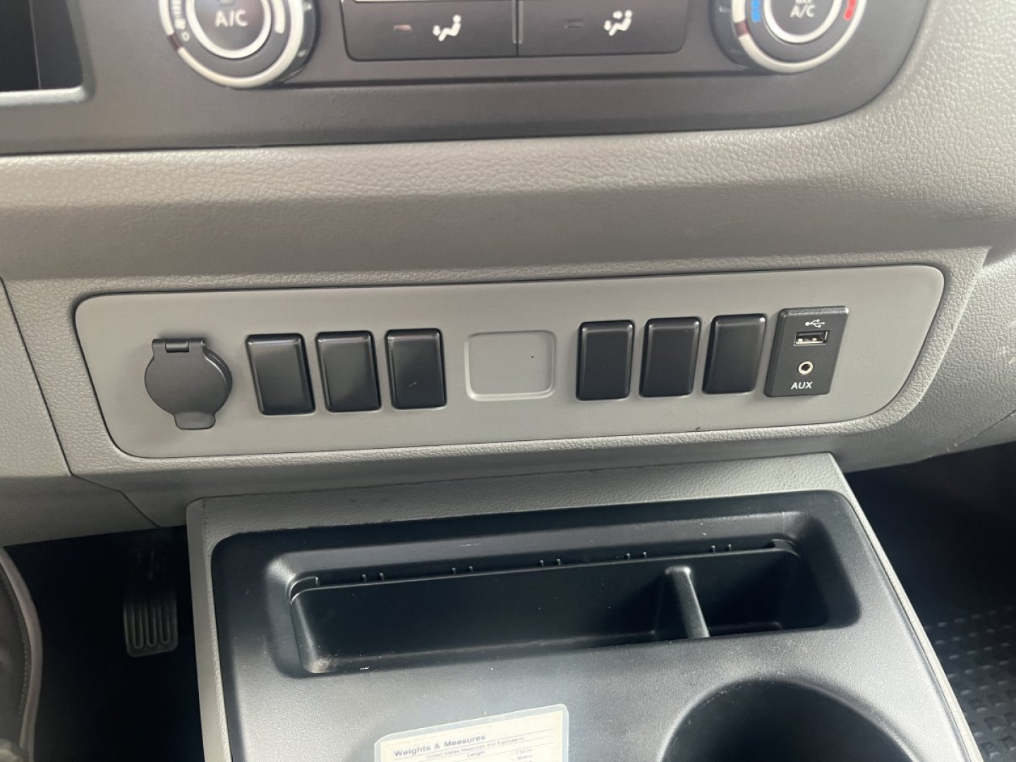 Dashboard area of a 2019 Nissan NV Cargo showing a row of switch panels and a small storage compartment beneath. Visible text includes vehicle controls and a power outlet cover with AUX label.