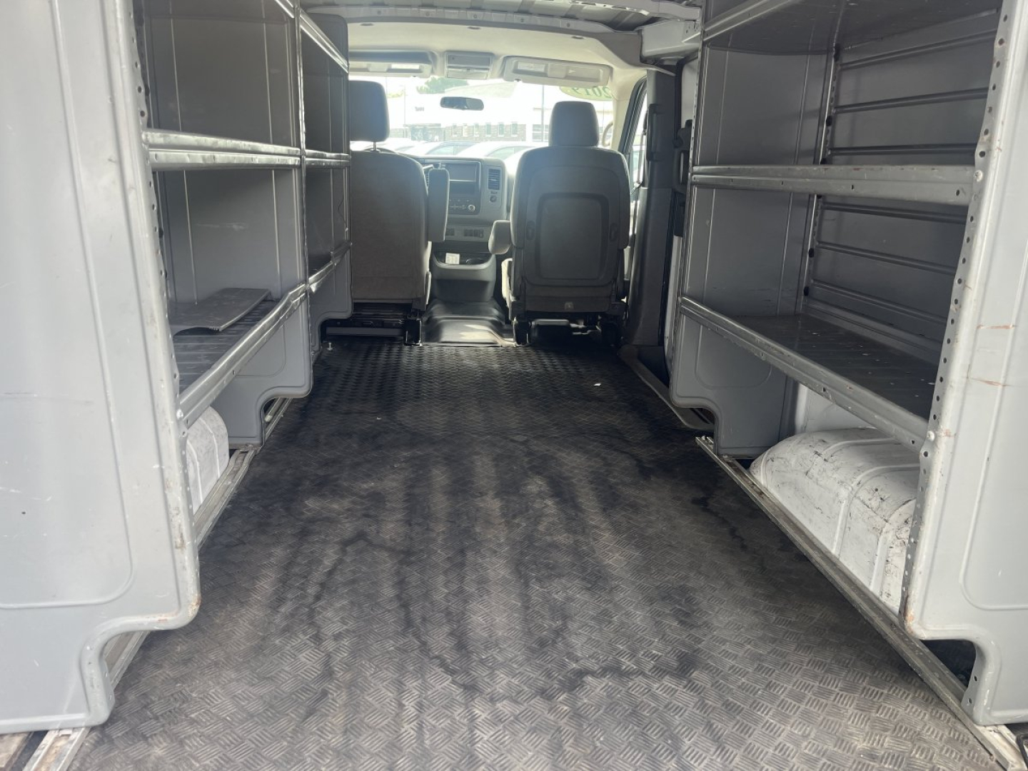 Van interior cargo area of a 2019 Nissan NV showing empty shelving along both sides and rubberized floor, front cabin visible from rear.