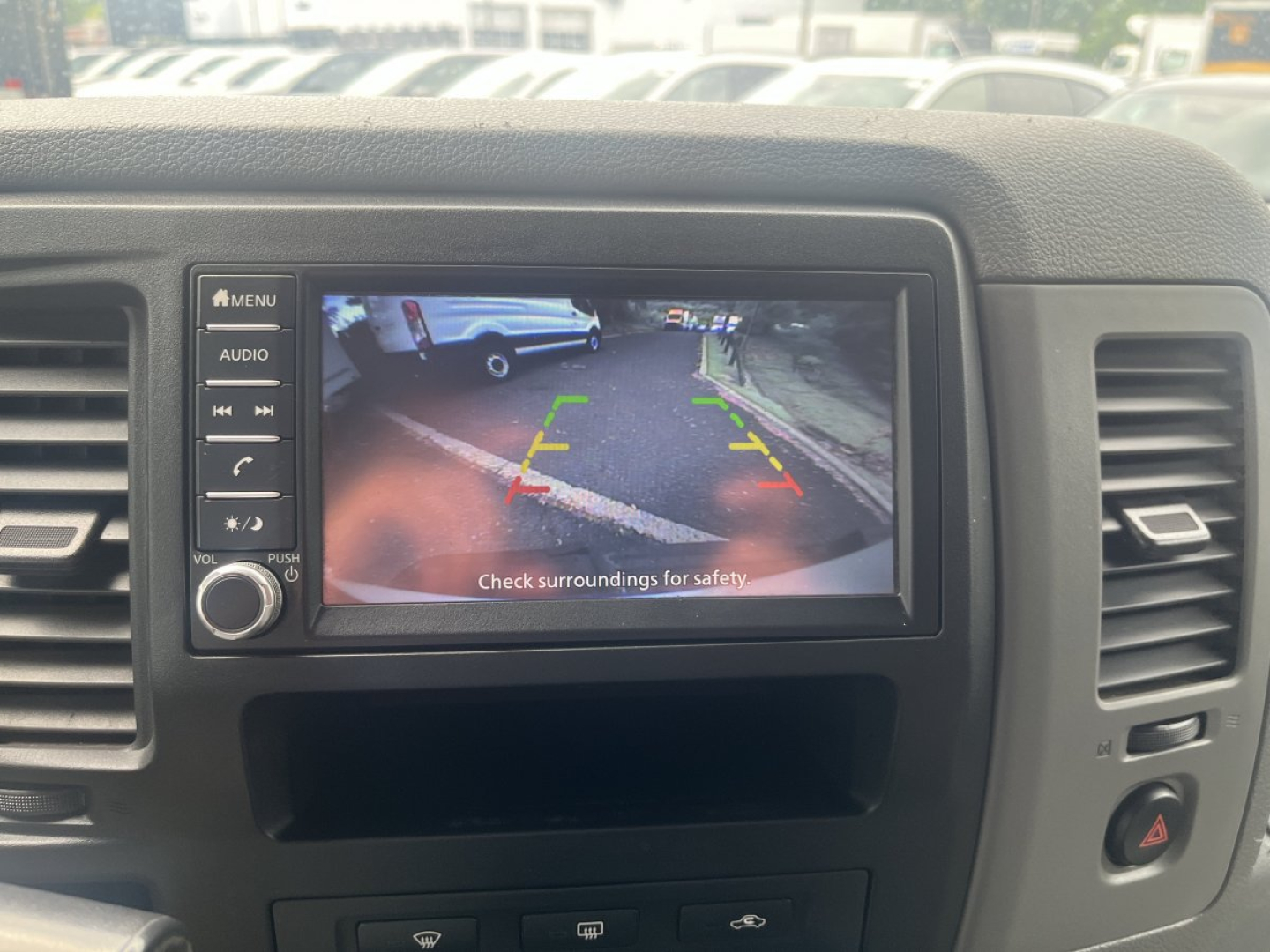 In-vehicle infotainment display showing backup camera view with parking guidelines in a Nissan NV Cargo cabin; surrounding dash controls visible.