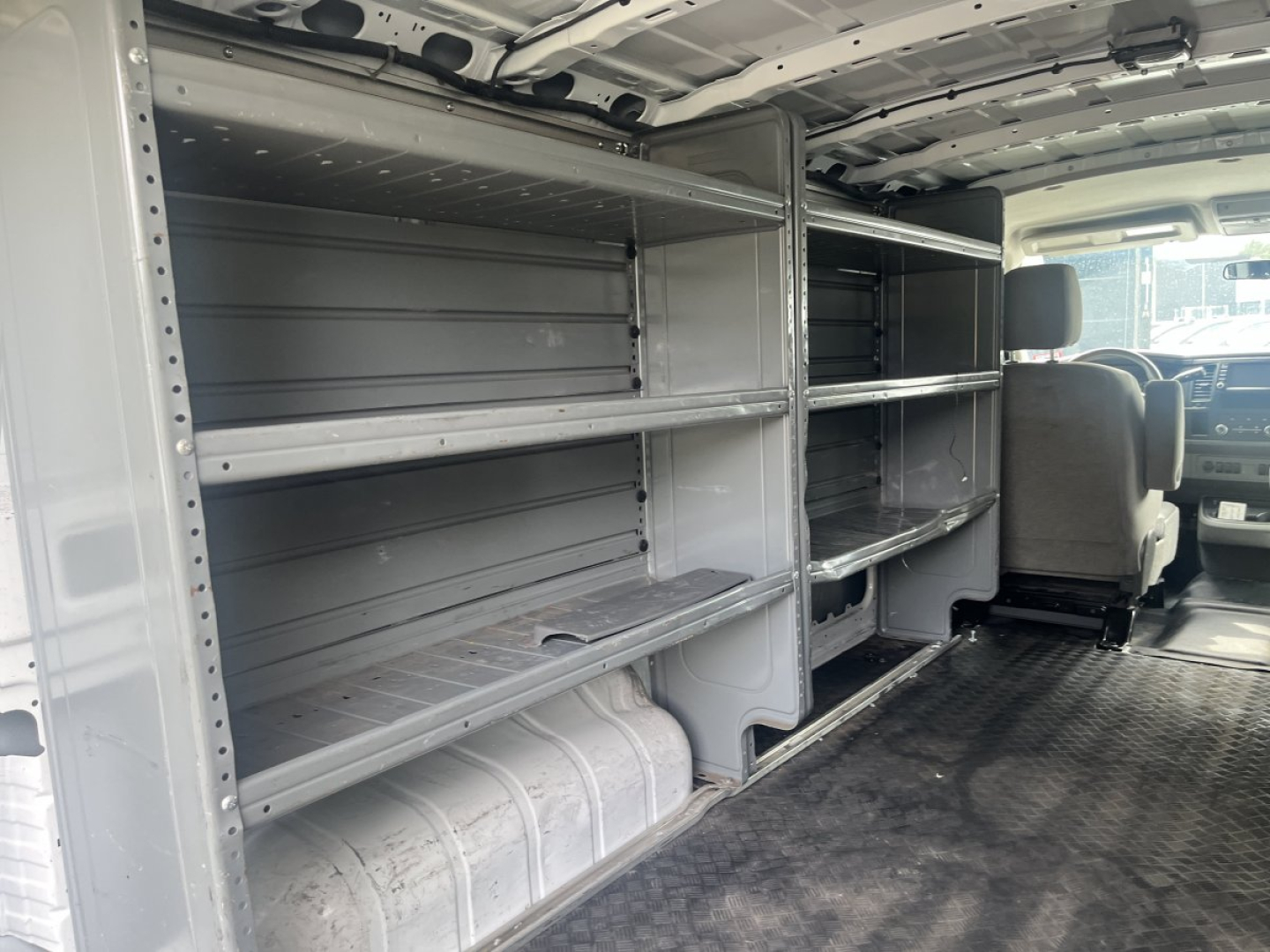Nissan NV Cargo van interior showing metal shelving and storage racks along both sides, with a gray rubberized floor and passenger cockpit visible in background.