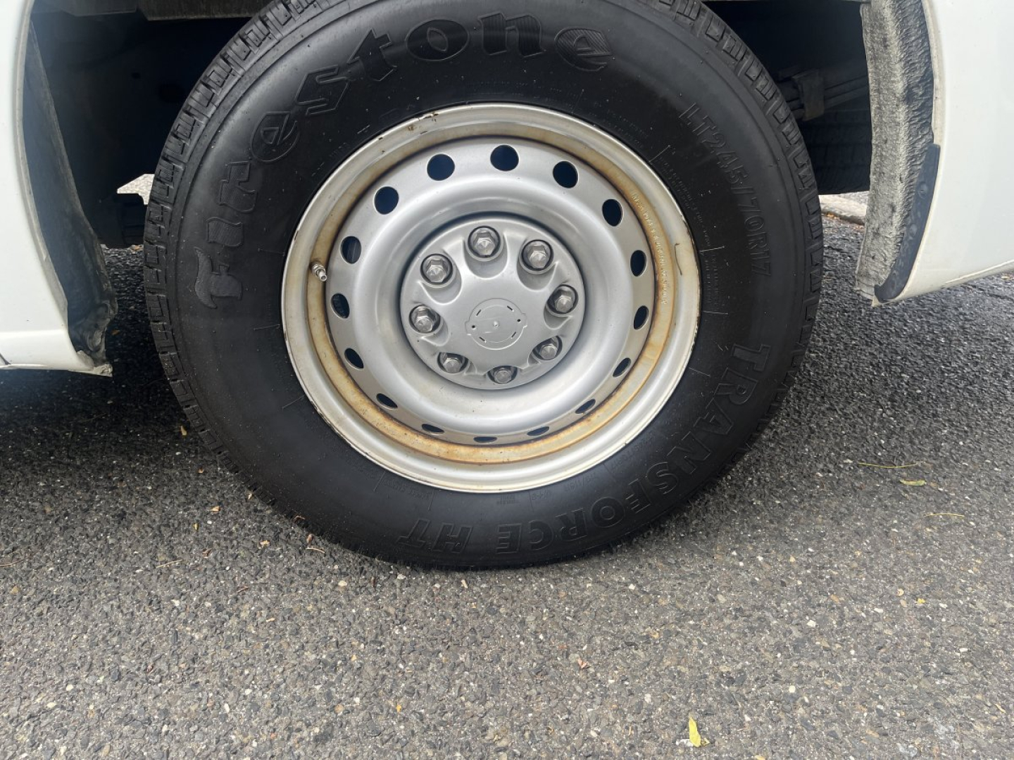 Wheel and tire close-up on a Nissan NV Cargo truck showing a steel wheel with multiple lug nuts and a Goodyear tire; side of vehicle and road surface visible.