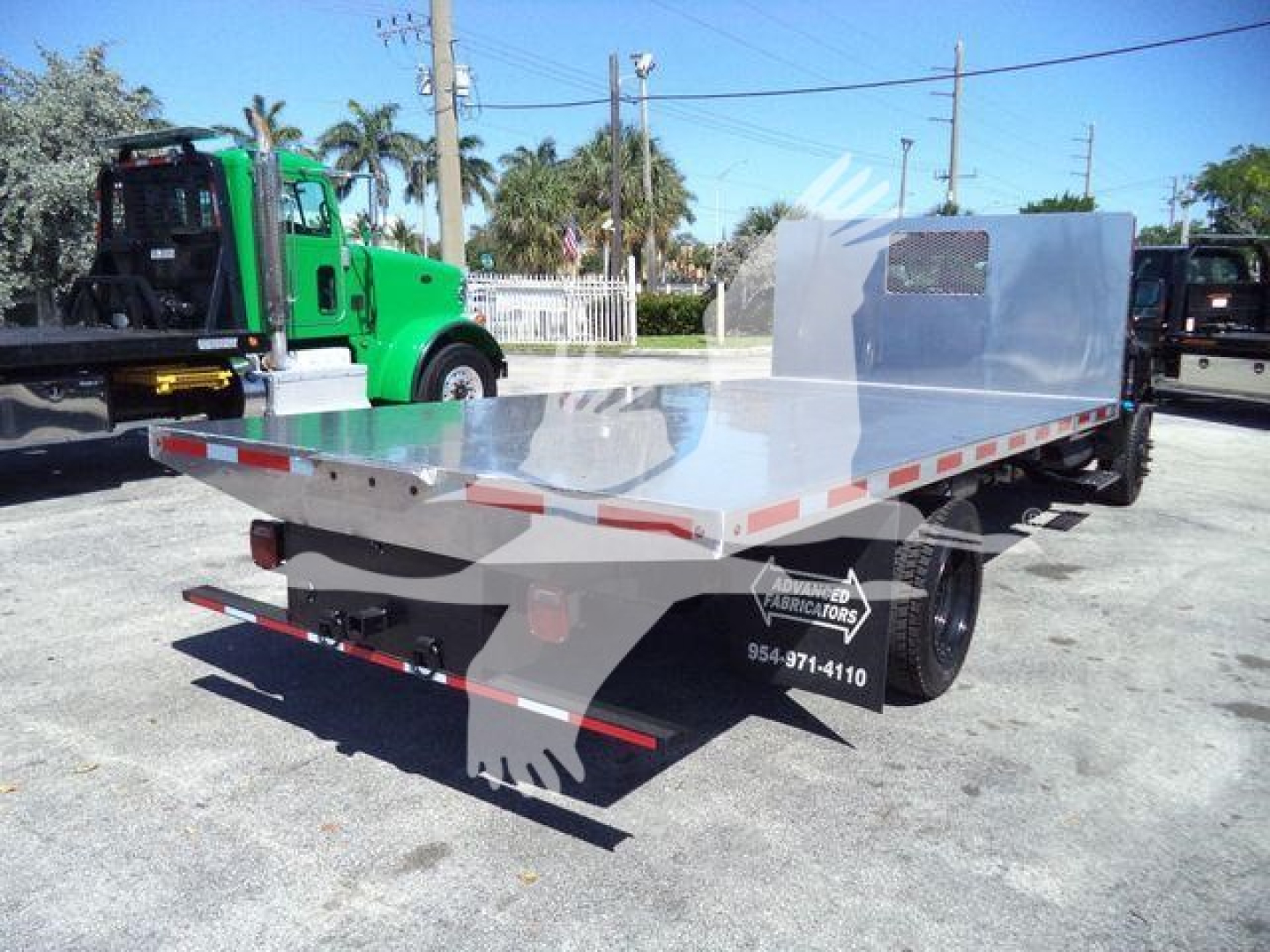 Flatbed truck bed on a 2023 Chevrolet Silverado 6500HD with aluminum flatbed and reflective tape, rear view showing mud flaps and wheel, green tow truck in background.