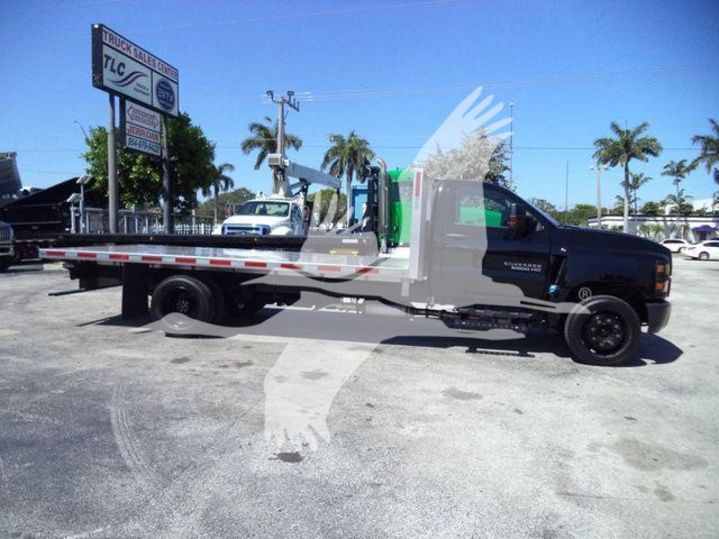 Flatbed tow truck side view showing Silverado 6500HD with long aluminum flatbed and truck cab, stock number 6500ALUMFLAT visible on sign in lot.