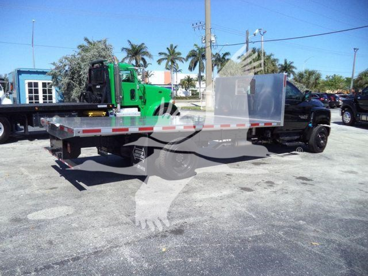 Flatbed tow truck with a green cab and aluminum flatbed, side rails, and a silver cab guard; visible asphalt surface and other trucks in background.