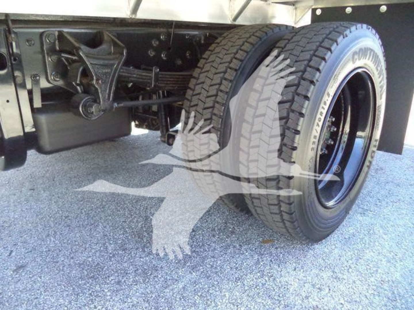 Truck rear axle area with dual rear wheels and flatbed frame; visible components include suspension linkage, leaf springs, and wheel tires.