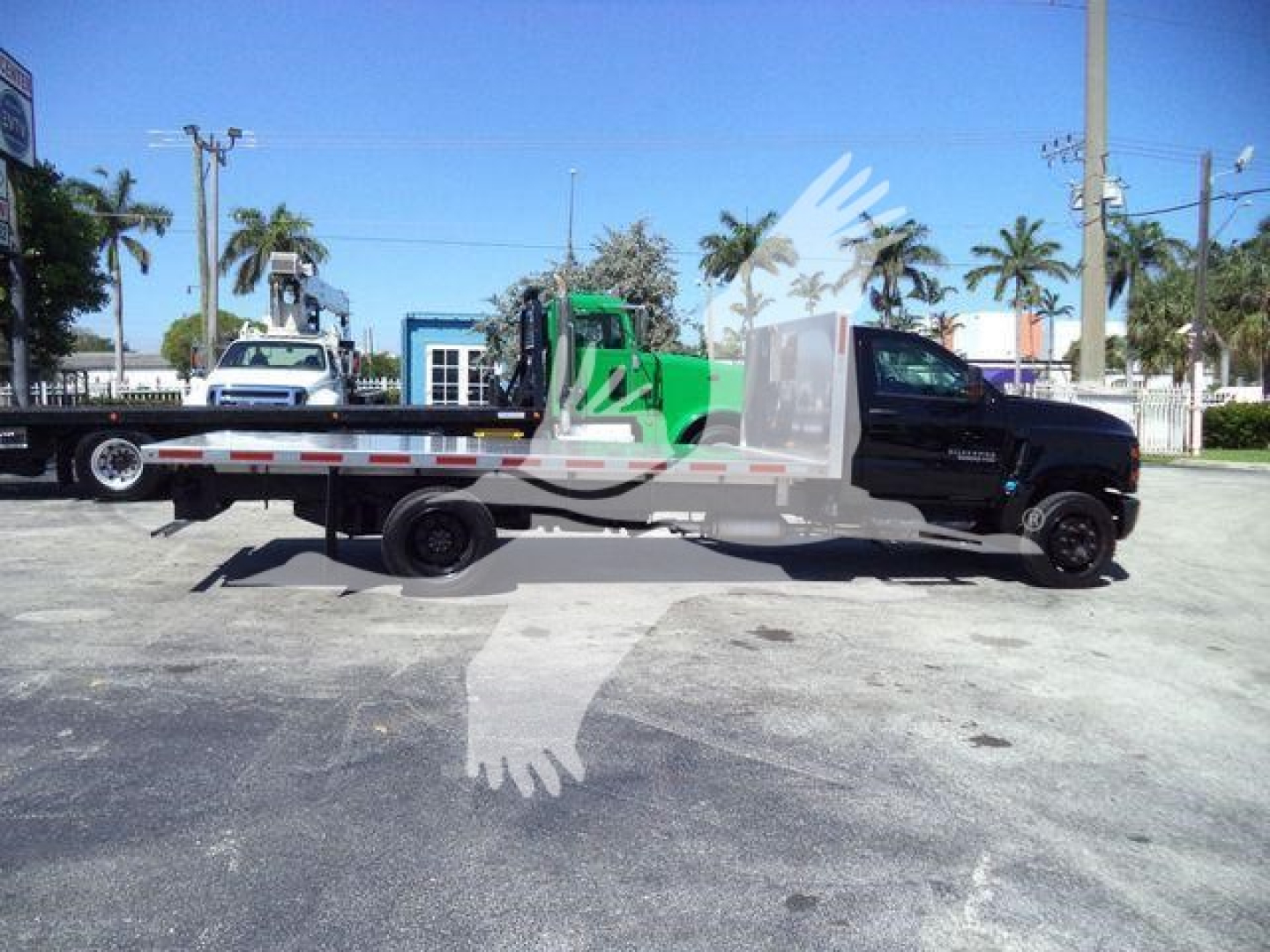 Flatbed tow truck side view showing 2023 Chevrolet Silverado 6500HD chassis with long aluminum flatbed and green crane attachment in lot.