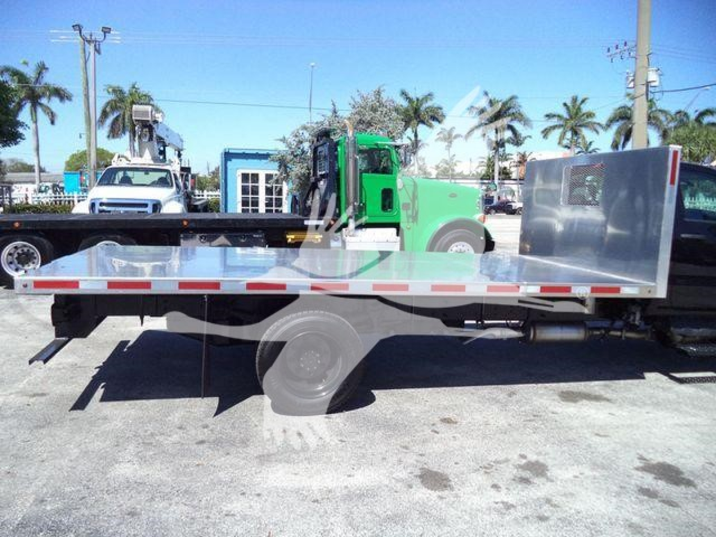 Flatbed truck with aluminum flatbed and green cab in background showing side view of Silverado 6500HD flatbed. Visible wheel, reflective tape, and metal side panel.