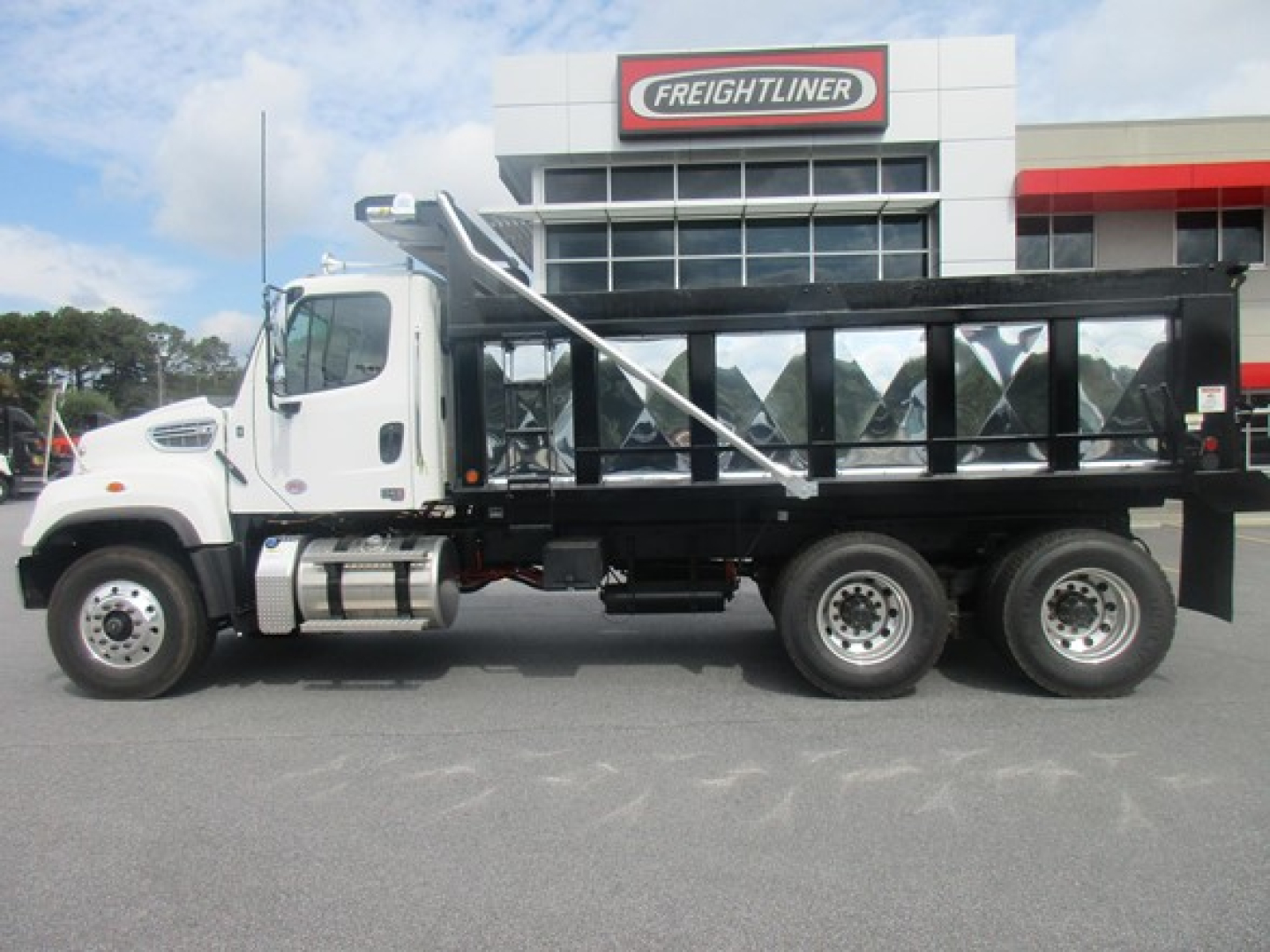 Freightliner 114SD dump truck side view with white cab and black dump bed, dealership building in background.