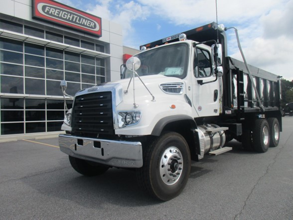 Freightliner 114SD dump truck in showroom lot with white cab and black dump body, Freightliner dealership in background.
