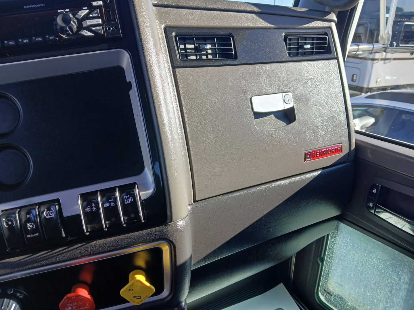 Dashboard area of a 2020 Kenworth T800 truck cab showing infotainment screen, control switches, and glovebox with Kenworth badge.