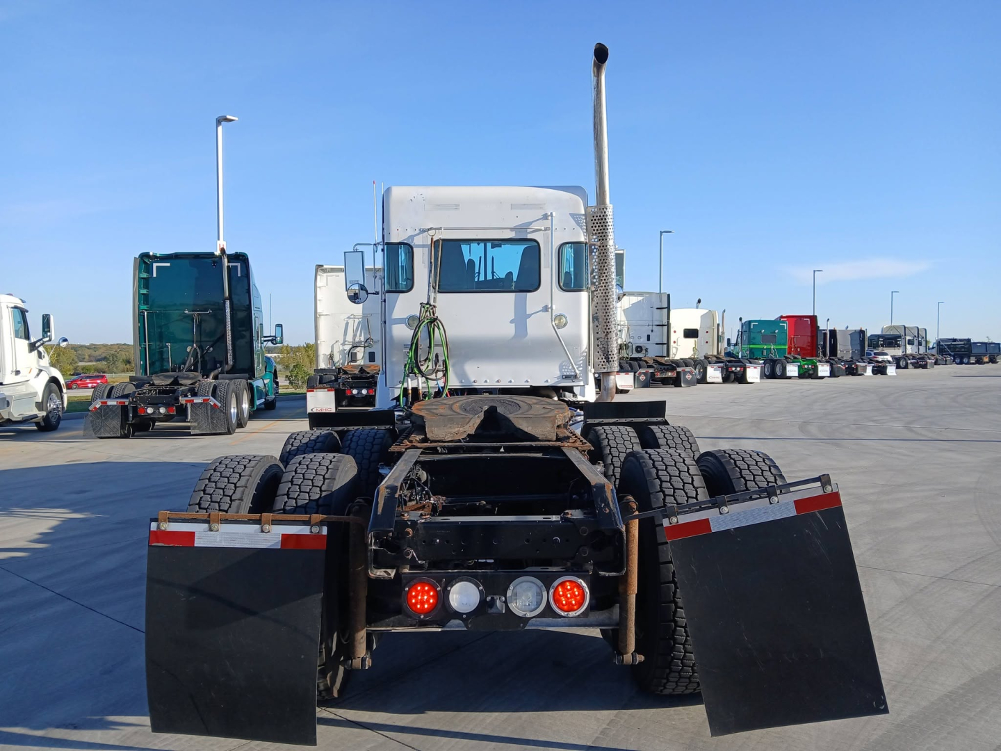 Kenworth truck tractor rear view with fifth wheel and mud flaps in a truck lot. Identifiable text not visible on components in this view.