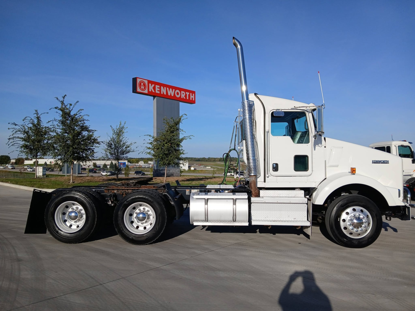 Kenworth T800 truck side view showing sleeper cab, dual rear axles, chrome exhaust stack, white paint, and Kenworth sign in background.