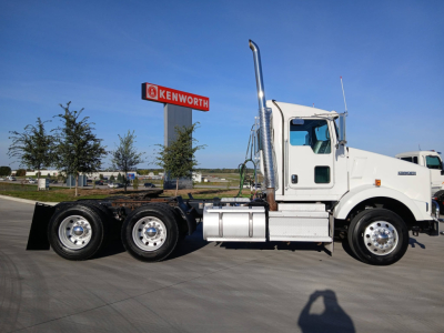 Kenworth T800 truck side view showing sleeper cab, dual rear axles, chrome exhaust stack, white paint, and Kenworth sign in background.