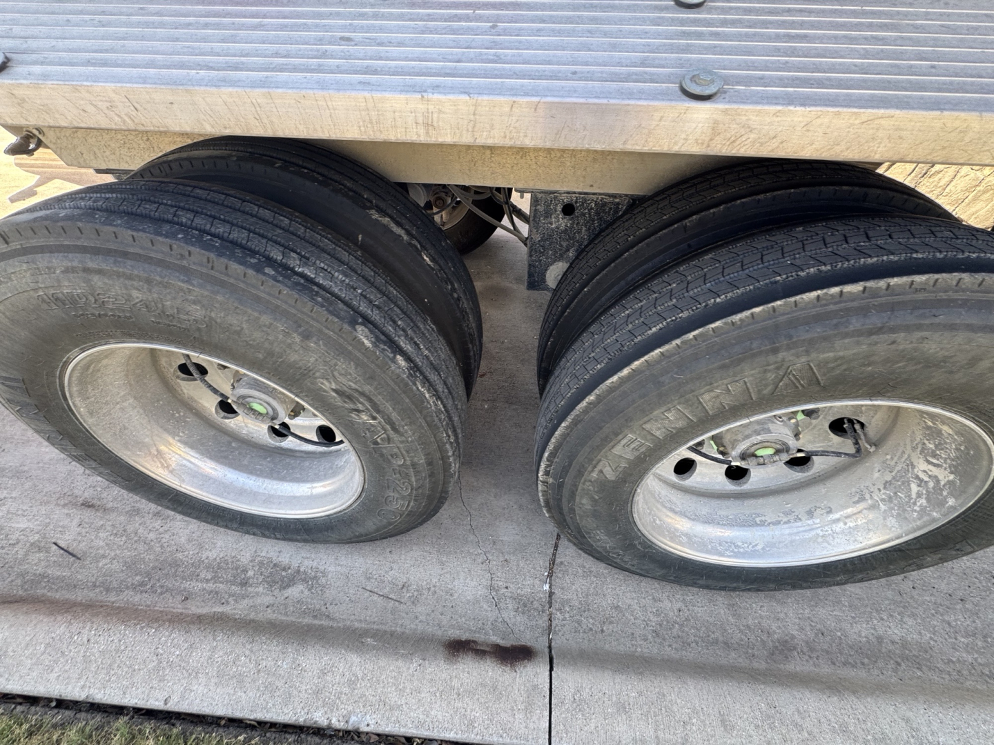 Dual rear trailer wheels and a portion of the aluminum deck showing two tires and rims under a dump trailer.
