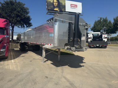 Trailer in a dealership lot showing a silver aluminum dump trailer with a gooseneck hitch and landing gear, side rails, and reflective tape along the edges.