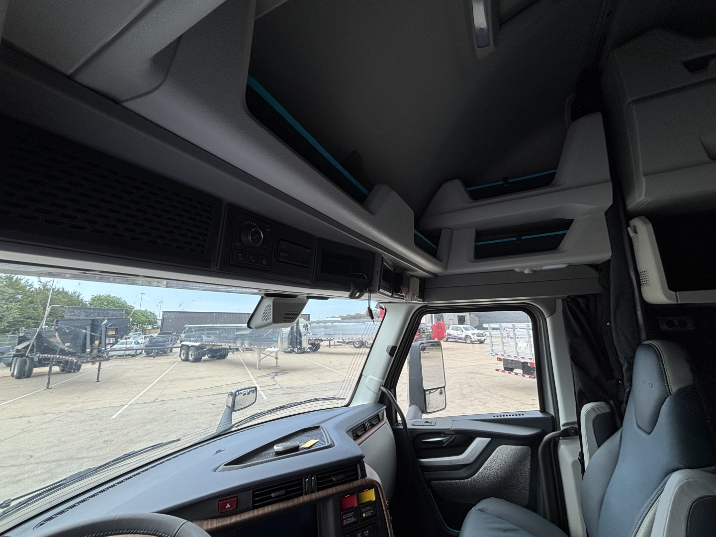 Cab interior of a 2027 Volvo VNL64T860. View shows upper storage compartments, dash area, steering wheel, and front windshield with trucks visible outside.