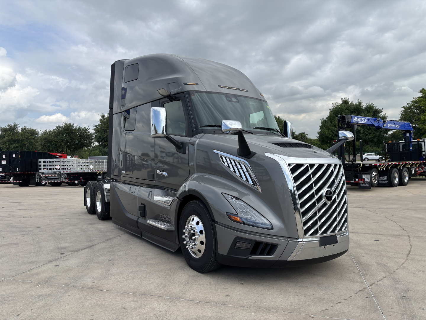 New 2027 Volvo VNL64T860 tractor-trailer front-left view with chrome grille and sleeper cab, grey exterior, stacked dual rear wheels, dealership lot background.