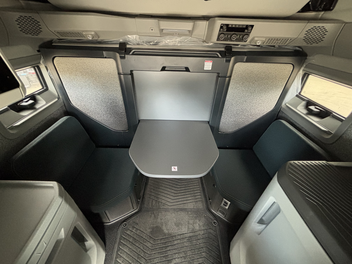 Truck cab interior showing sleeper area with table and seating; overhead control panel and window side panels visible.