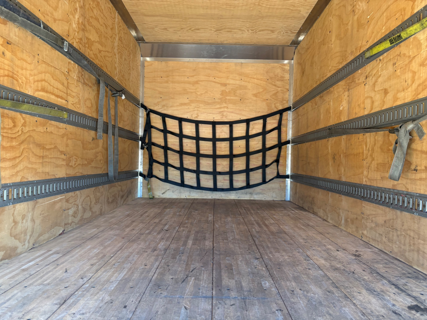 Box truck cargo area with wooden floor and side walls, black cargo net at back, metal E-track rails along walls, tie-down hooks, empty interior.